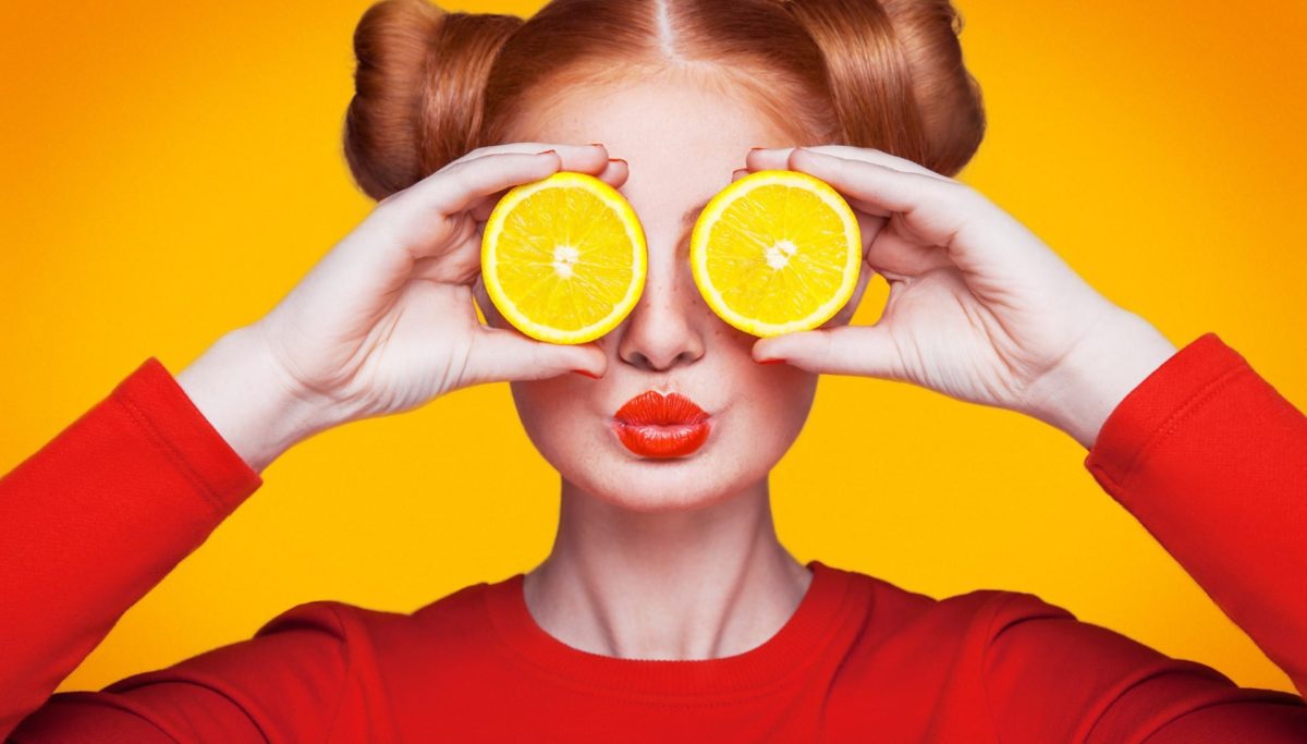 Young beautiful funny fashion model with lemon slice on orange background. with makeup and hairstyle and freckles. holding lemon between eyes with kiss.