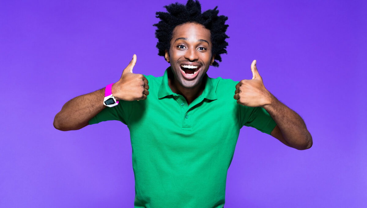 Portrait of happy afro american young man wearing green polo shirt, laughing at camera with thumbs up. Studio shot on violet background.