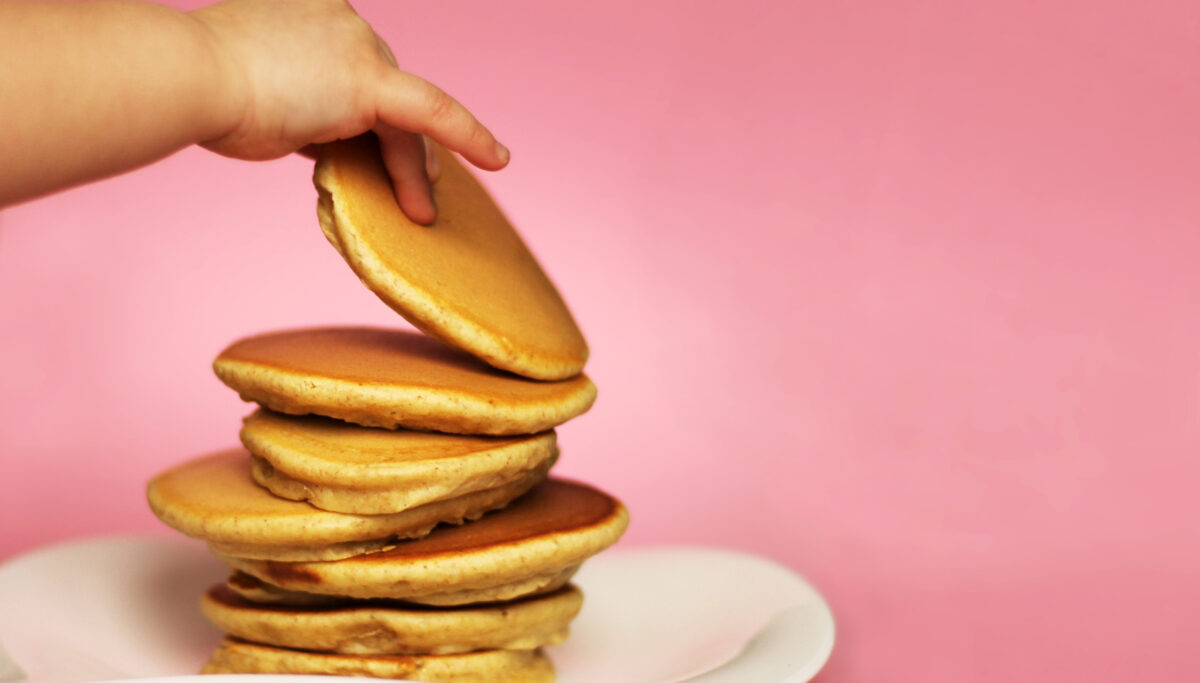 A child takes a pancake from a stack, pink background. American delicious pancakes. Close up, copy space