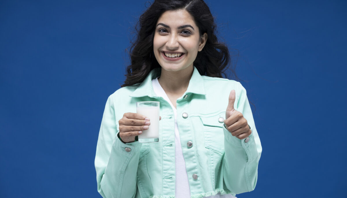Woman holding a glass of milk in one hand and her thumb up with the other hand on a blue background.