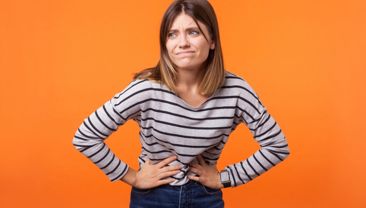 Portrait of unhappy bloated woman with brown hair in long sleeve striped shirt standing, holding her belly with hands, stomach cramps or period pain. indoor studio shot isolated on orange background